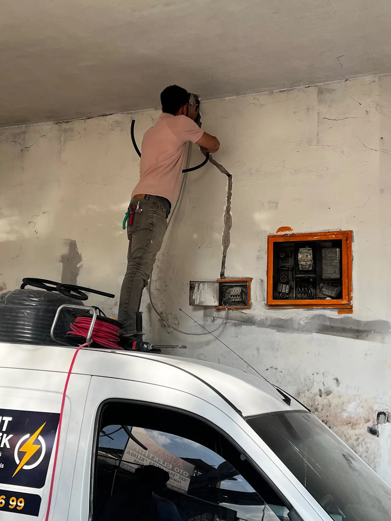 Turkut Elektrik ustası duvardaki sayaç panosuna yeni hat bağlarken; önde firmanın aracı